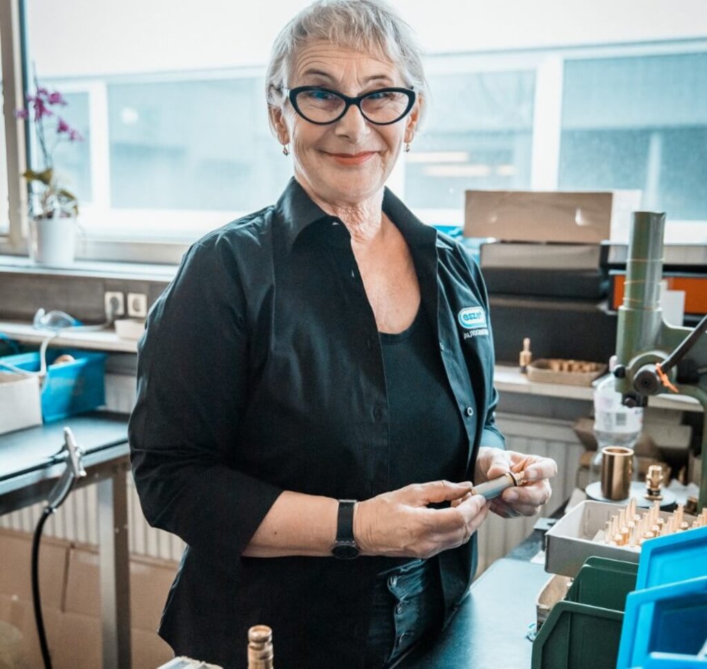 Mitarbeiterin hält ein Bauteil in der Hand und arbeitet an einem Produktionsplatz.