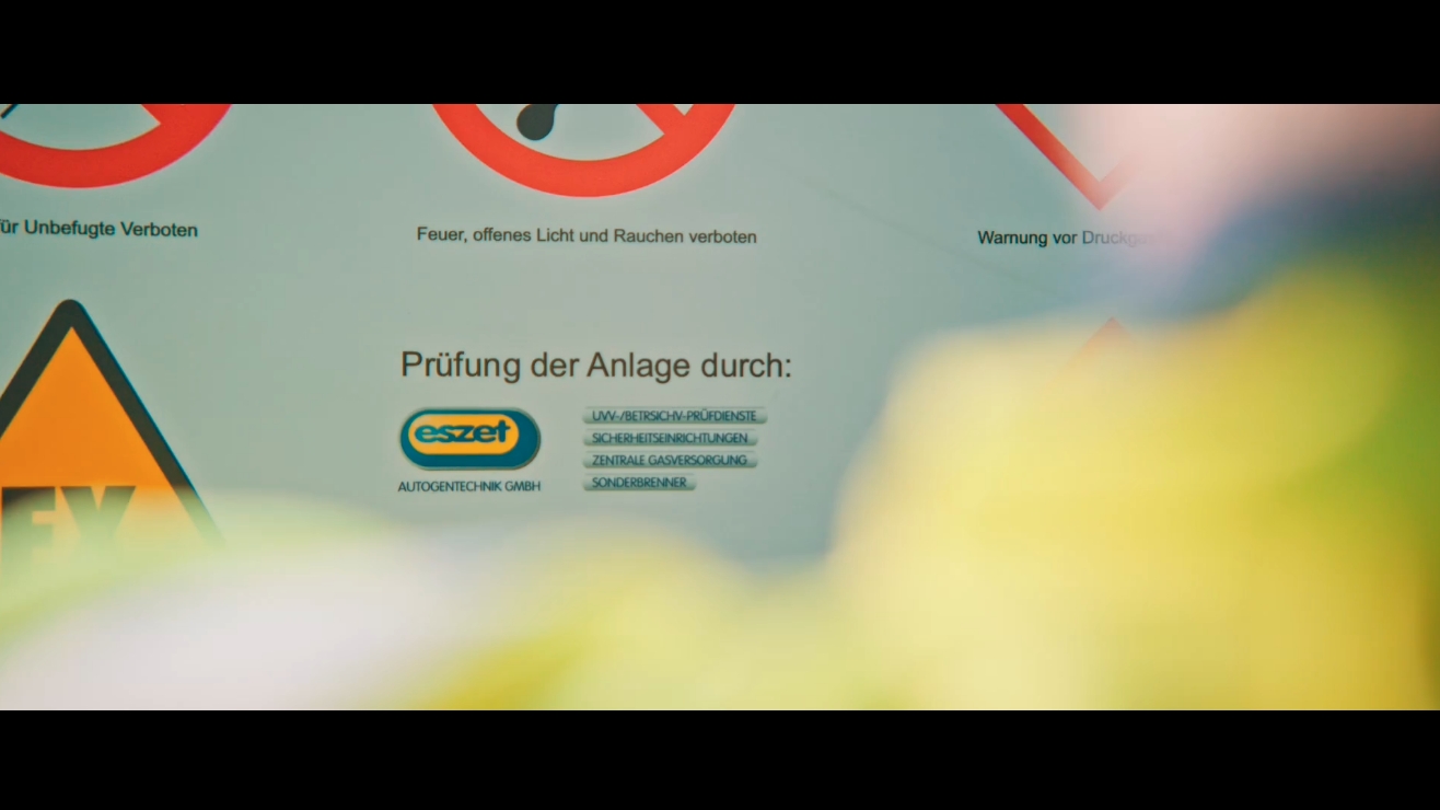 Hinweisschild mit dem Text „Prüfung der Anlage durch Eszet“ und Sicherheitsymbolen im Hintergrund.